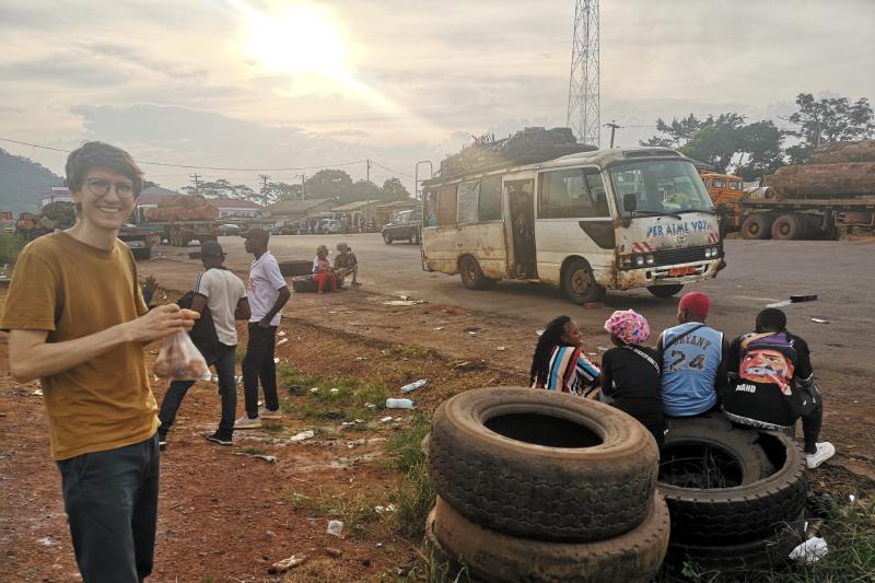 Bus am Straßenrand im Vordergrund sitzen Menschen auf Reifen, im Hintergrund ein LKW mit riesiegen Baumstämmen.
